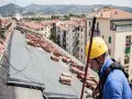 Operaio con casco giallo lavora sul tetto, tra pile di tegole, per riparare infiltrazioni.