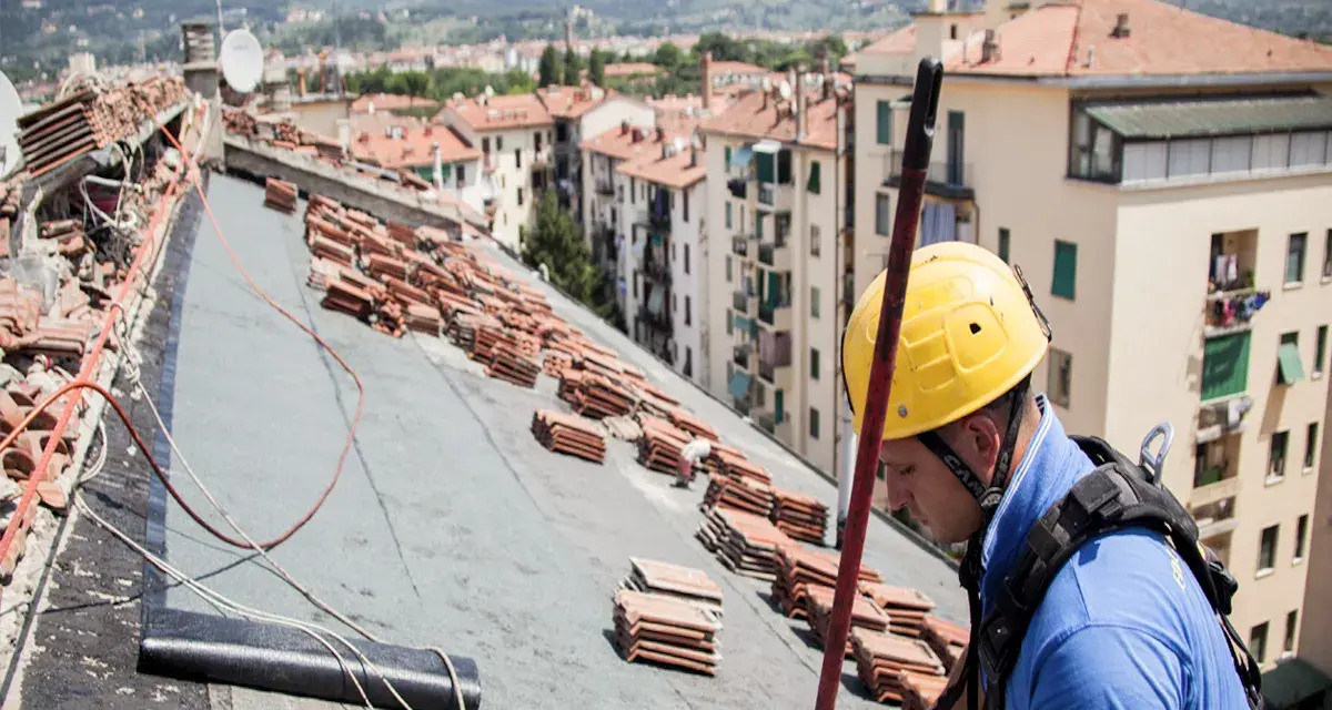 Operaio con casco giallo lavora sul tetto, tra pile di tegole, per riparare infiltrazioni.