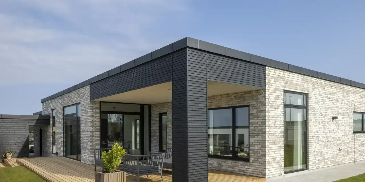 Casa moderna con mattoni chiari, ampie vetrate e **tetti piani moderni**. Un deck in legno circonda parte dell'abitazione.