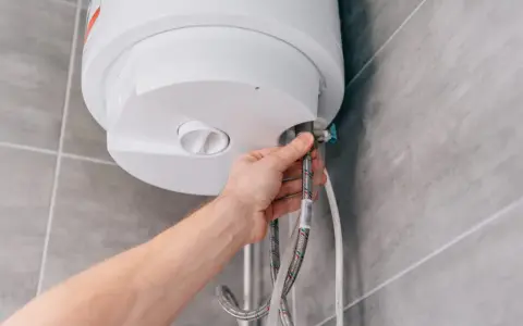 Mano che controlla la valvola scaldabagno perde. Tubi flessibili collegati a un boiler bianco su sfondo piastrellato.