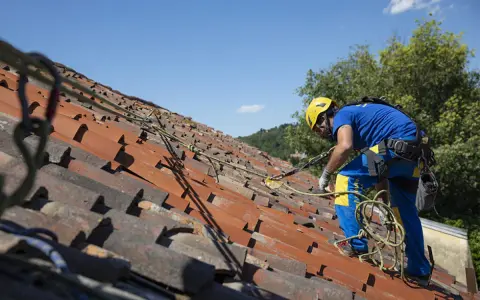 Operaio con imbracatura di sicurezza lavora sul tetto per rifare il tetto, assicurato da cavi e moschettoni.