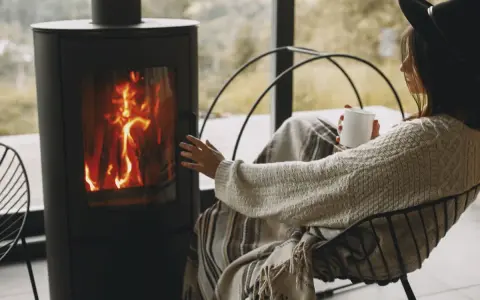 Donna avvolta in una coperta, con una tazza in mano, si scalda vicino a una stufa a legna, un modo accogliente per riscaldare la casa senza termosifoni.