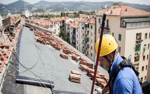 Operaio con casco giallo lavora sul tetto, tra pile di tegole, per riparare infiltrazioni.