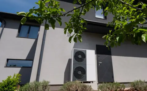 Unità esterna di una pompa di calore, moderna e discreta, installata accanto a una porta su una casa grigia.