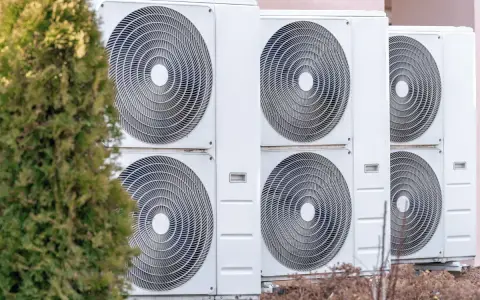 Unità esterne di climatizzatori, parte di un sistema di riscaldamento elettrico, allineate accanto a un albero verde.