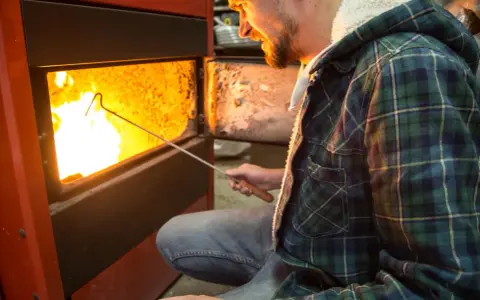 Uomo con camicia a quadri verde accende un fuoco in uno scaldabagno a legna, mostrando come funziona.