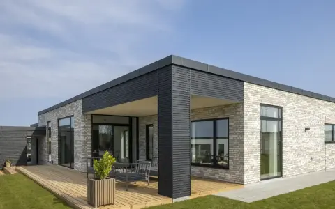 Casa moderna con mattoni chiari, ampie vetrate e **tetti piani moderni**. Un deck in legno circonda parte dell'abitazione.