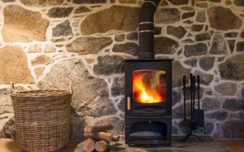 Stufa a pellet accesa su muro in pietra. Con cesto di vimini e attrezzi per camino. Potrei installare una stufa a pellet da solo.