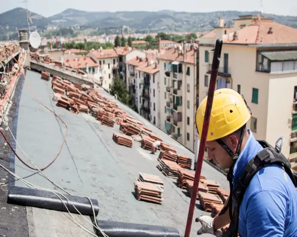 Operaio con casco giallo lavora sul tetto, tra pile di tegole, per riparare infiltrazioni.