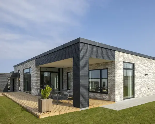 Casa moderna con mattoni chiari, ampie vetrate e **tetti piani moderni**. Un deck in legno circonda parte dell'abitazione.
