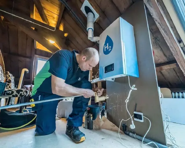Installazione di caldaie a idrogeno in un sottotetto. Un tecnico collega tubi a un'unità bianca con il simbolo H2.