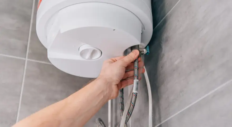 Mano che controlla la valvola scaldabagno perde. Tubi flessibili collegati a un boiler bianco su sfondo piastrellato.