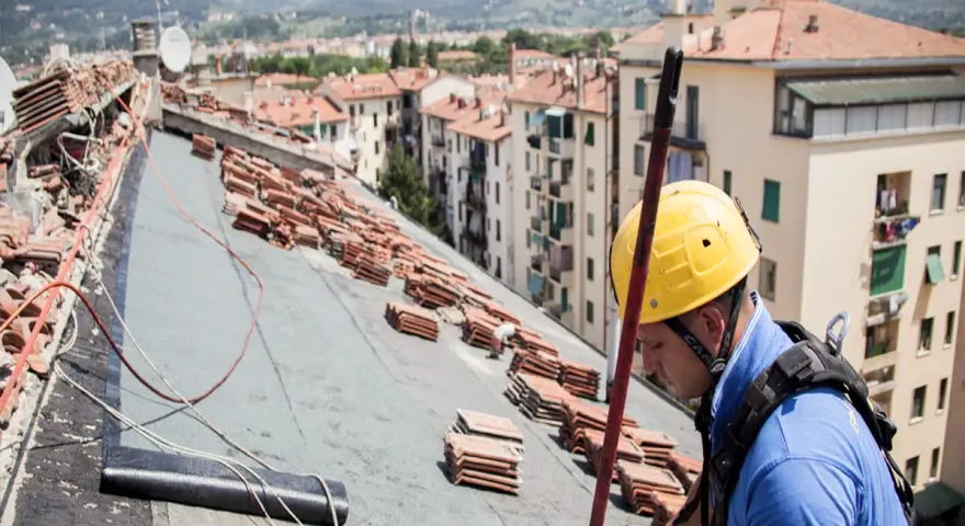 Operaio con casco giallo lavora sul tetto, tra pile di tegole, per riparare infiltrazioni.