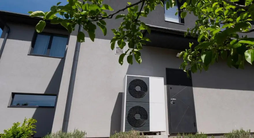 Unità esterna di una pompa di calore, moderna e discreta, installata accanto a una porta su una casa grigia.