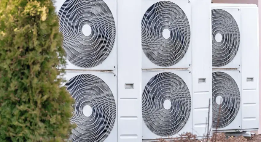Unità esterne di climatizzatori, parte di un sistema di riscaldamento elettrico, allineate accanto a un albero verde.