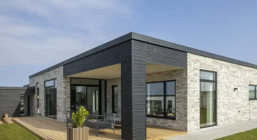 Casa moderna con mattoni chiari, ampie vetrate e **tetti piani moderni**. Un deck in legno circonda parte dell'abitazione.