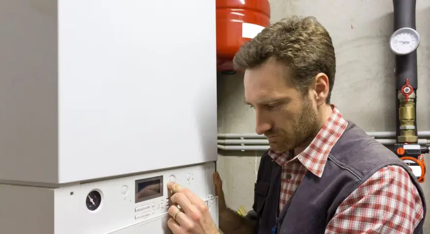Un tecnico con una camicia a quadri e un gilet grigio regola un pannello di controllo di una caldaia bianca. Guida nella scelta della caldaia.