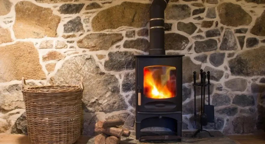 Stufa a pellet accesa su muro in pietra. Con cesto di vimini e attrezzi per camino. Potrei installare una stufa a pellet da solo.