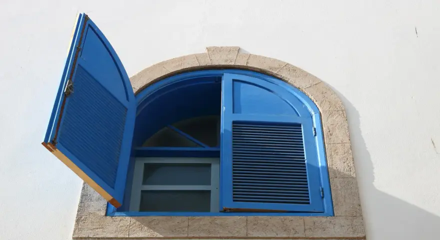 Finestre aperte o chiuse d'estate, con persiane blu su muro bianco. La luce del sole crea ombre nette.