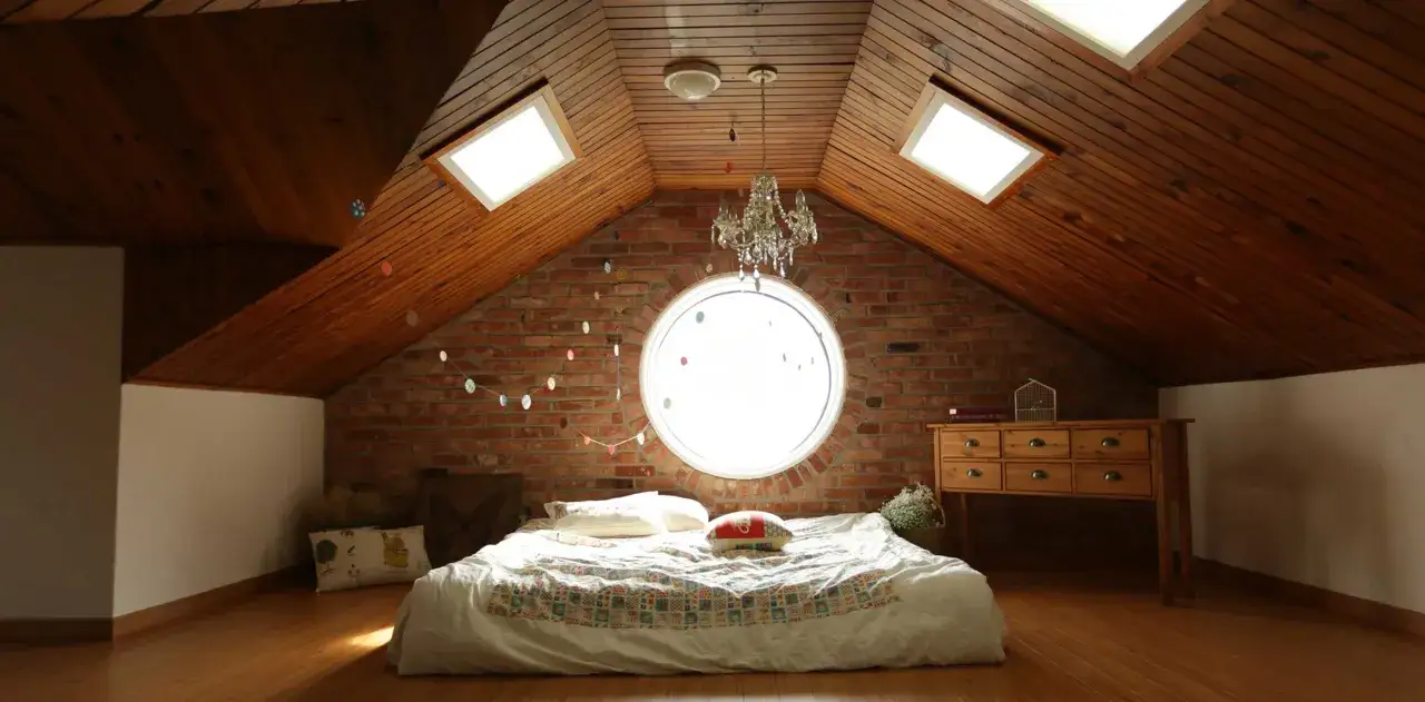 Camera puntata verso un letto in mansarda, con il soffitto in legno e la sua evidente pendenza tetto. Finestre sul tetto e un obl&ograve; circolare illuminano la stanza.